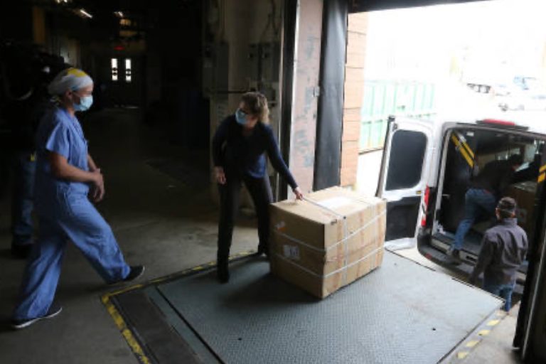Two individuals in blue scrubs loading boxes into a van, possibly for medical supplies transportation.